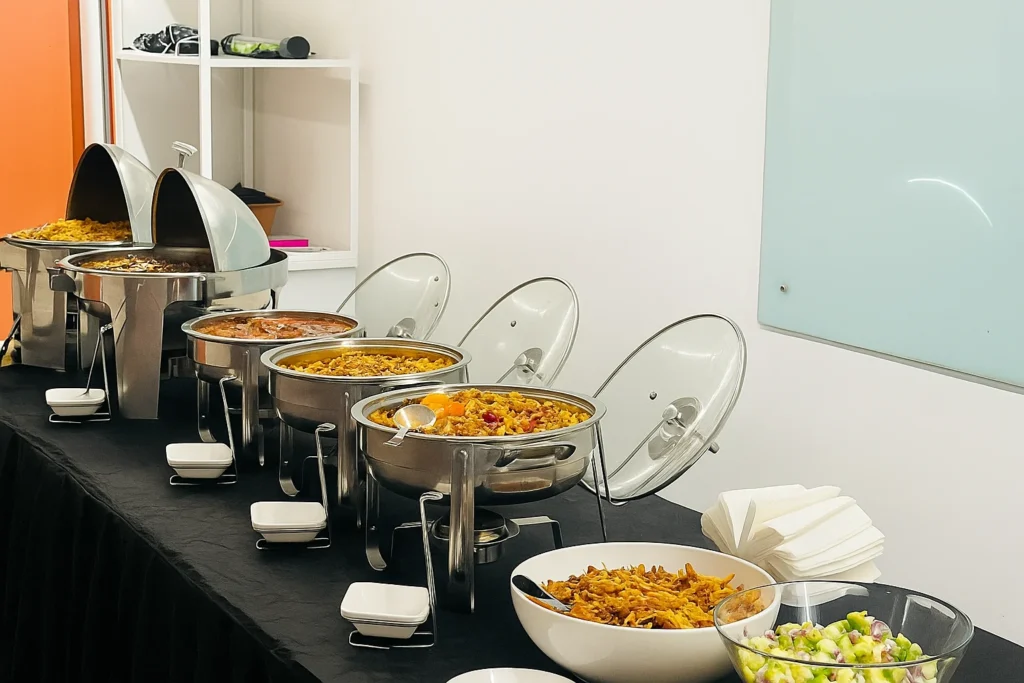 Buffet setup in a modern indoor venue with stainless steel chafing dishes, rice and pasta dishes, fresh salad bowls, and neatly arranged napkins — representing professional corporate catering by SAB Hospitality.